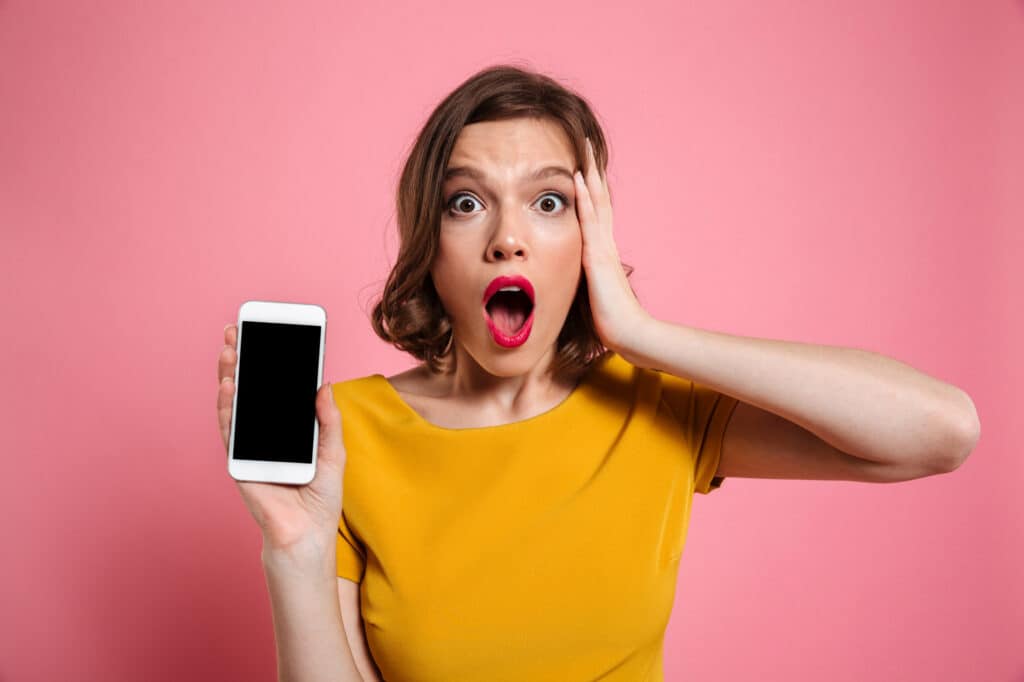 Eine Frau in einem gelben T-Shirt hält sich die linke Hand an den Kopf. mit der rechten Hand hält sie ihr Mobiltelefon in die Kamera.