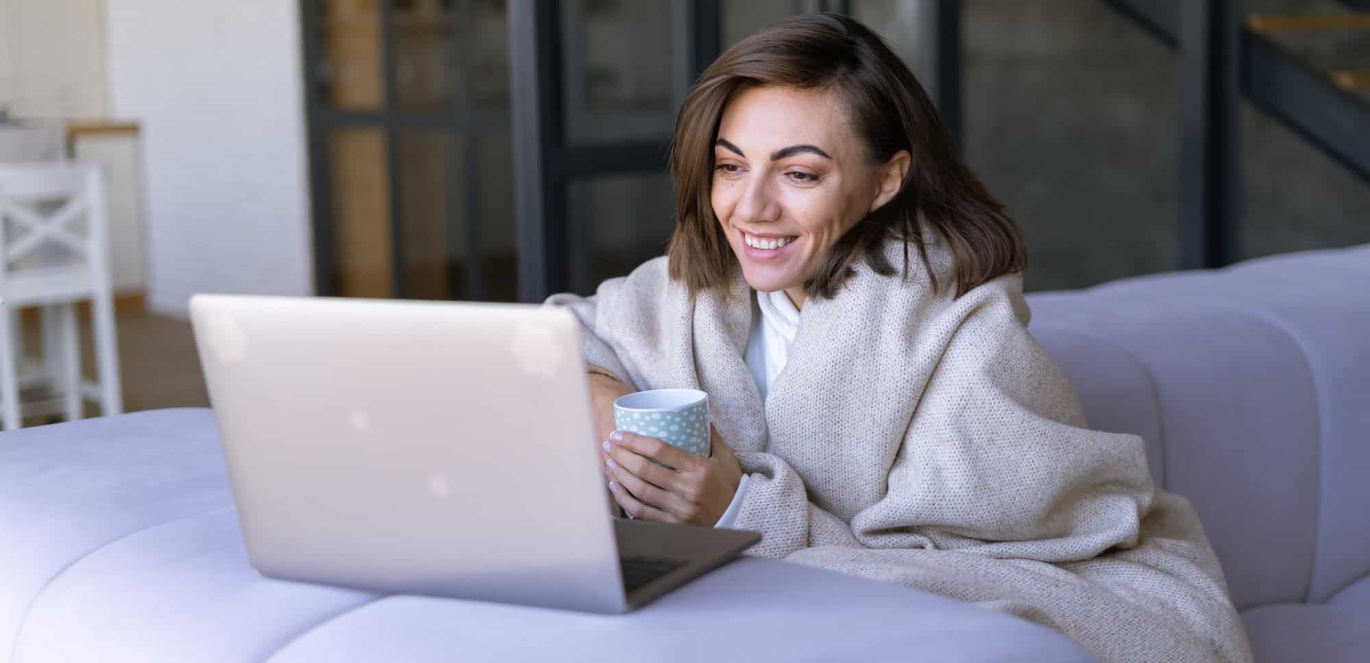 Eine junge Frau sitzt zuhause auf der Couch und lächelt in ihren Laptop. In der Hand hält sie eine Tasse mit Tee.