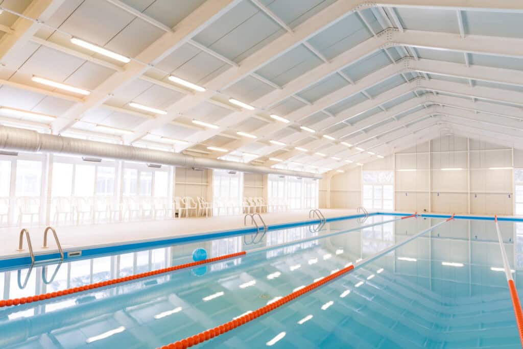 Das Bild zeigt ein leeres Hallenbad mit roten Schwimmbahnen. Die Sonne schient durch die großen Fenster herein.