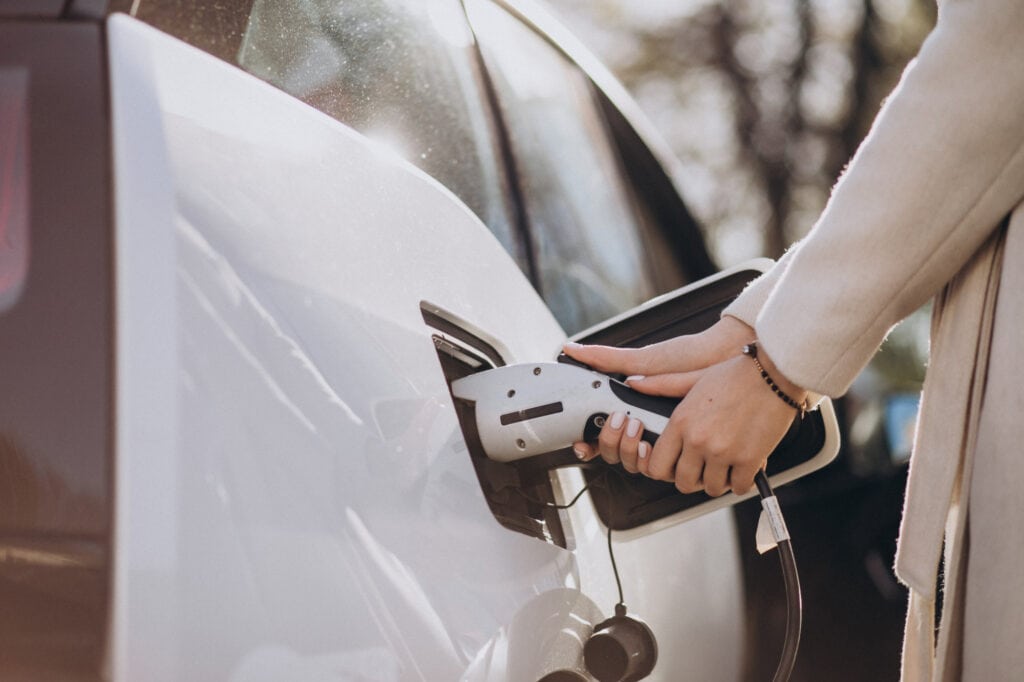 Es ist eine Frau in einem beigen Mantel zu sehen, die ein weißes Auto mittels einer Ladestation für Elektro-Autos lädt.