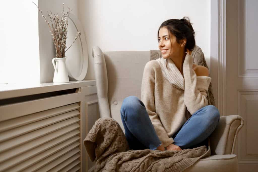 Eine lächelnde Dame sitzt mit Jeans und Pullover auf einem beigen Sessel an einem Fenster. An der Wand unterhalt des Fensters ist ein weißer Heizkörper verbaut.