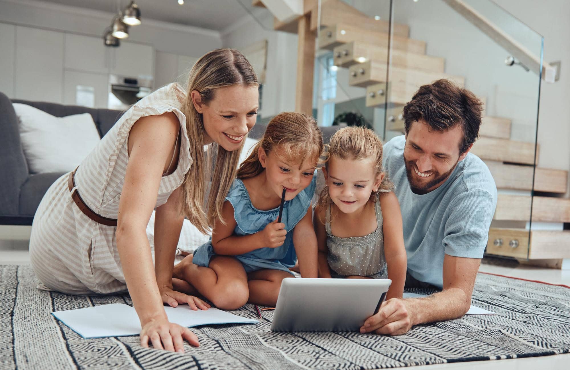 Das Bild zeigt eine Familie (Mann, Frau und zwei Kinder), die auf dem heimischen Boden im Wohnzimmer liegt und auf ein Tablet schaut.